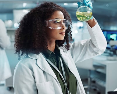 Scientist wearing safety goggles examining a flask with green liquid in a laboratory.