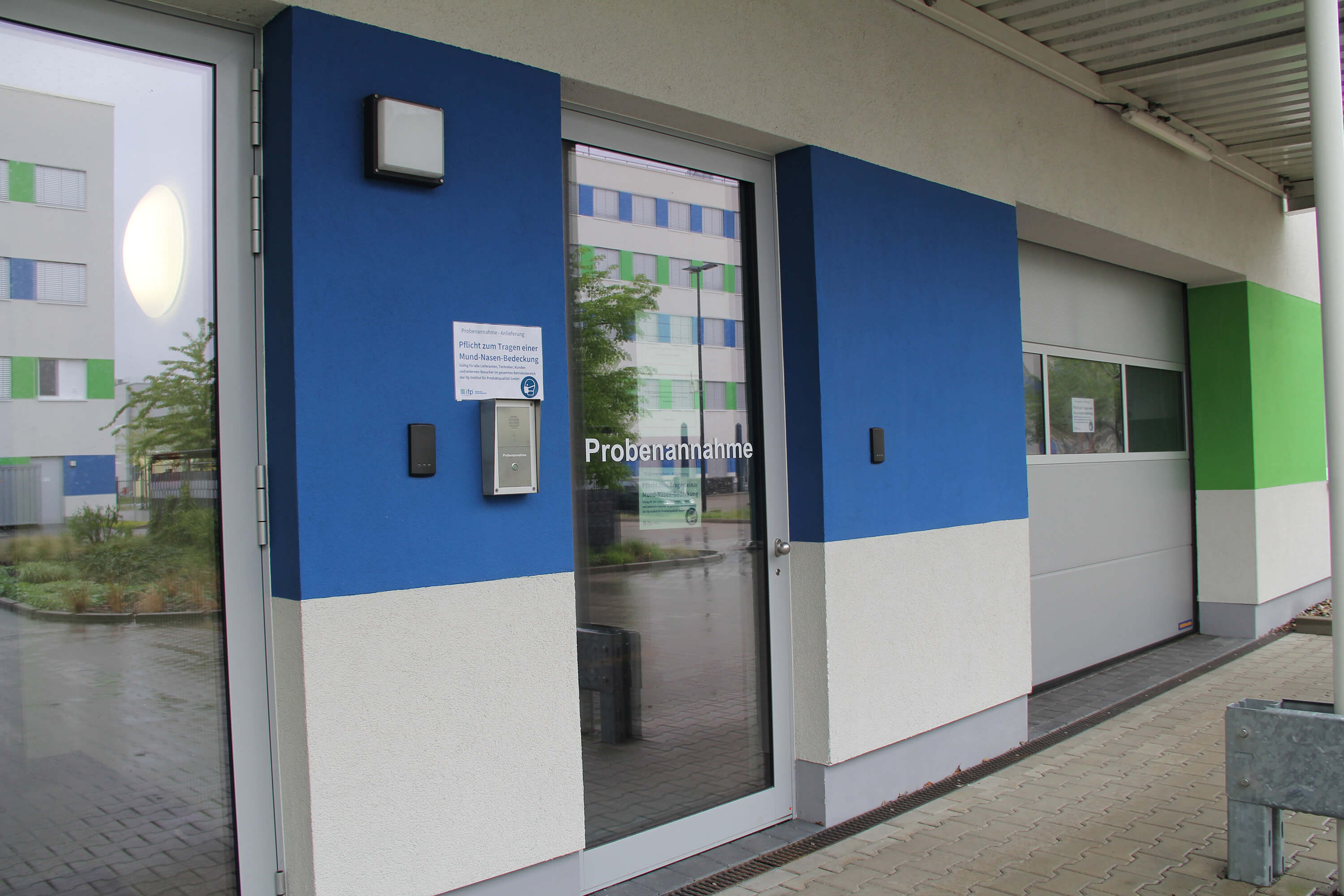 Exterior view of a laboratory or facility entrance with a glass door labeled ‘Probenannahme,’ indicating the sample collection area.