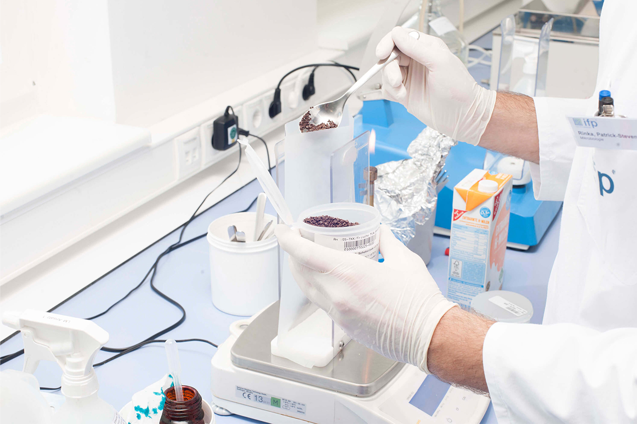 Scientist weighing granular sample material on a laboratory scale.