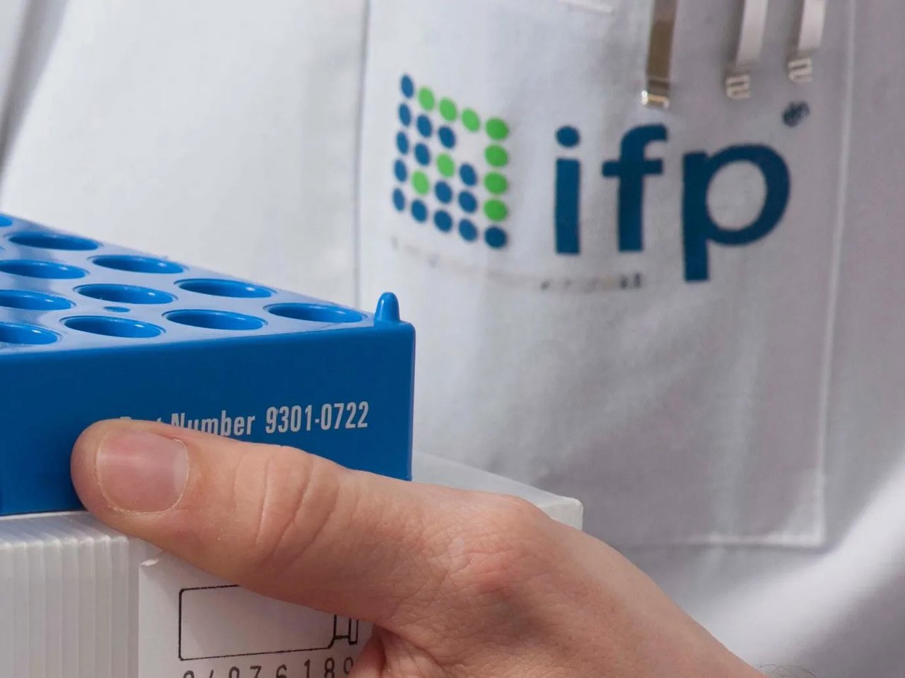 Close-up of a hand holding a blue laboratory sample rack with test tubes, with the ifplabs logo visible on the employee’s lab coat.