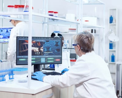 A senior scientist works in a pharmaceutical laboratory while a research team is visible in the background.