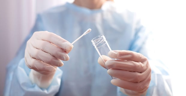 A gloved healthcare worker holds a test tube and cotton swab in a close-up view.