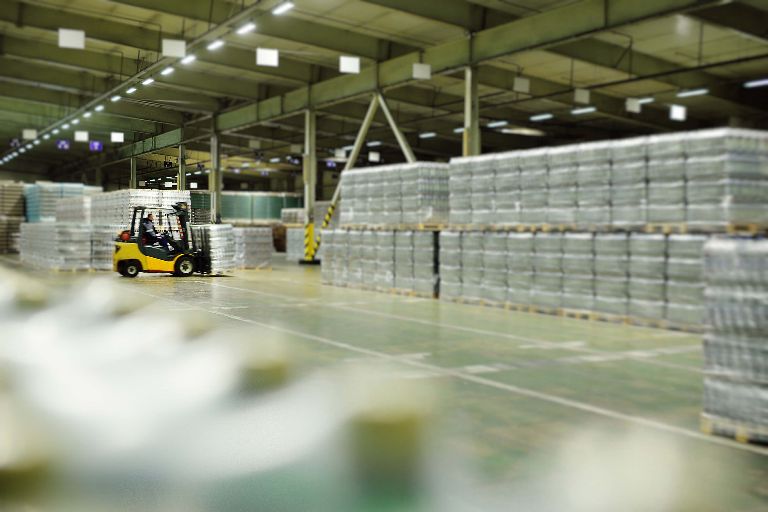 A yellow loader moves through a large beverage warehouse filled with plastic bottles.