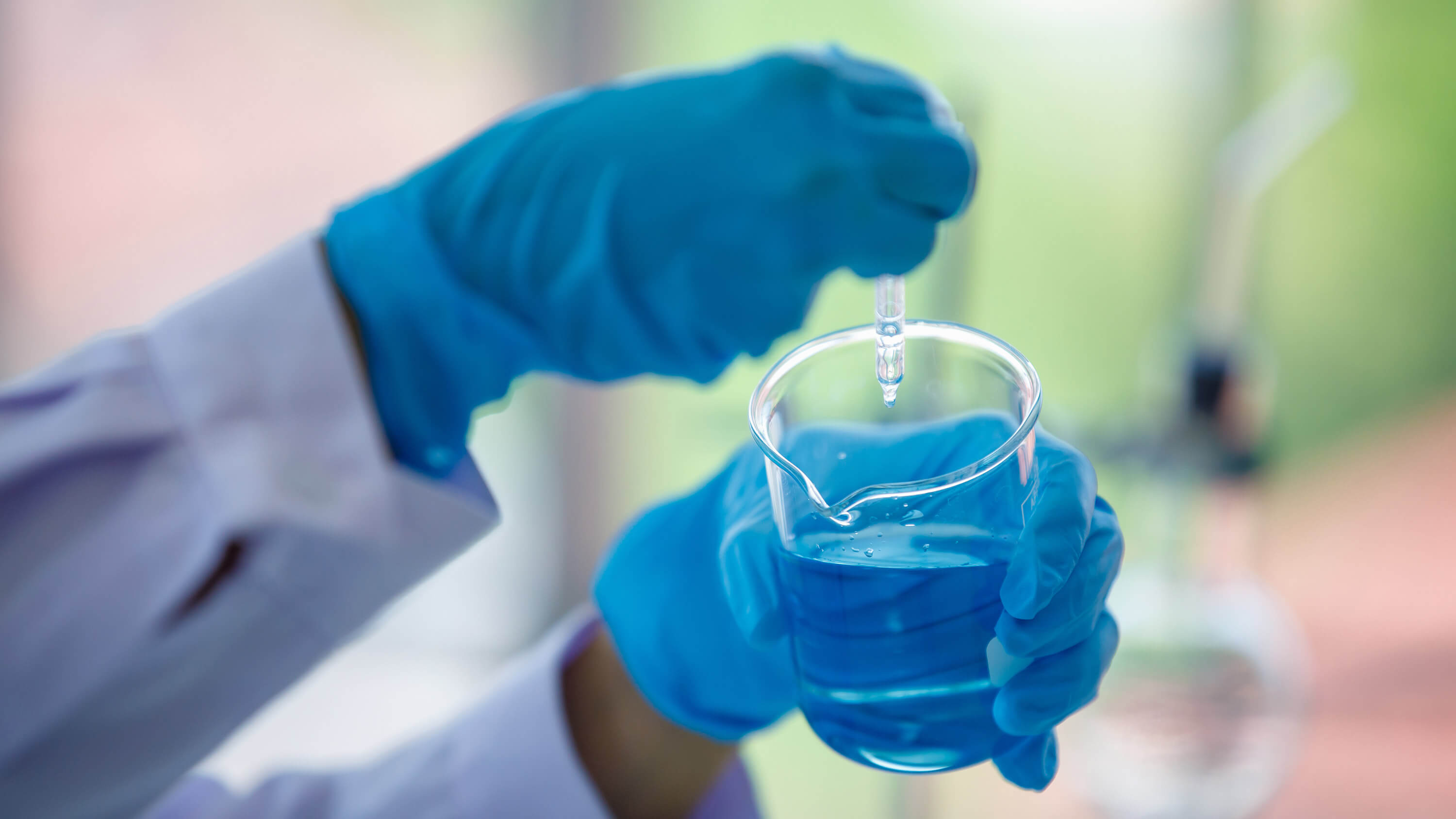 A gloved hand holds a laboratory beaker filled with blue liquid in a close-up view.