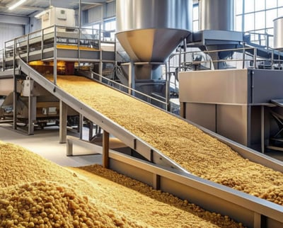An industrial facility processes large quantities of feed ingredients with conveyors, metal silos, and piles of pellet-like material.