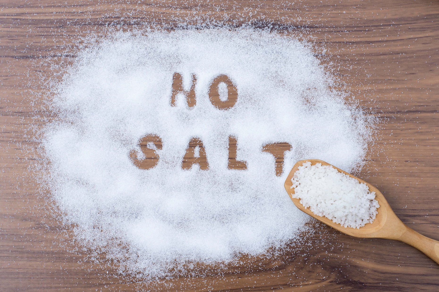 Wooden scoop with sea salt and the words no salt written in salt grains on a wooden table.