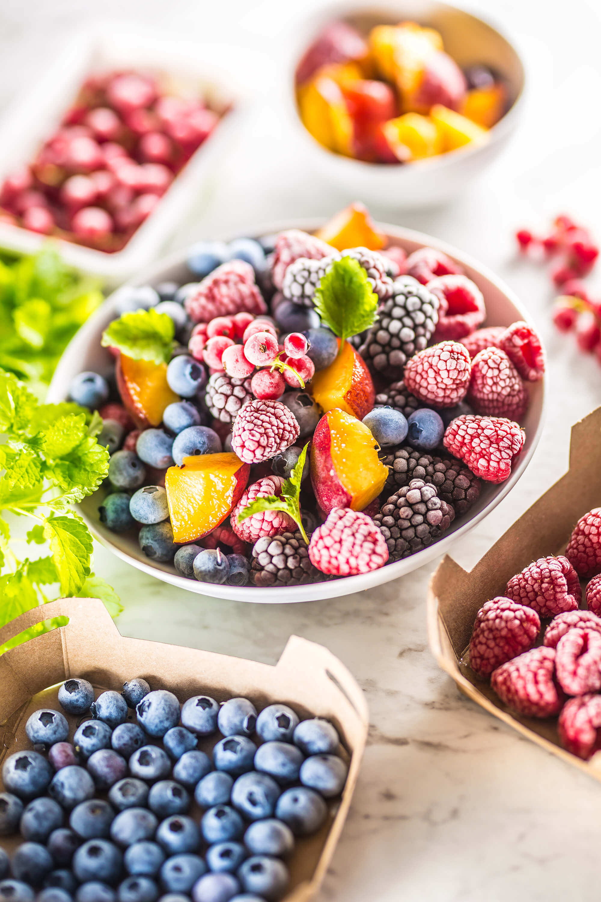 Frozen fruits including blueberries, blackberries, raspberries, red currants, peach slices, and lemon balm are arranged on a table.