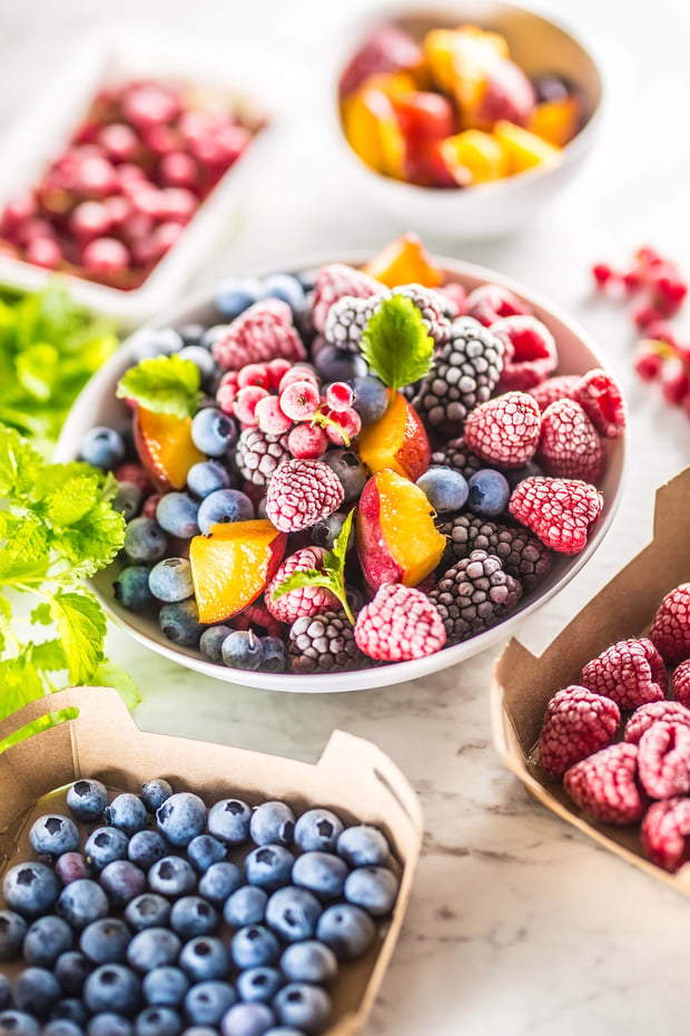 Frozen fruits including blueberries, blackberries, raspberries, red currants, peach slices, and lemon balm are arranged on a table.