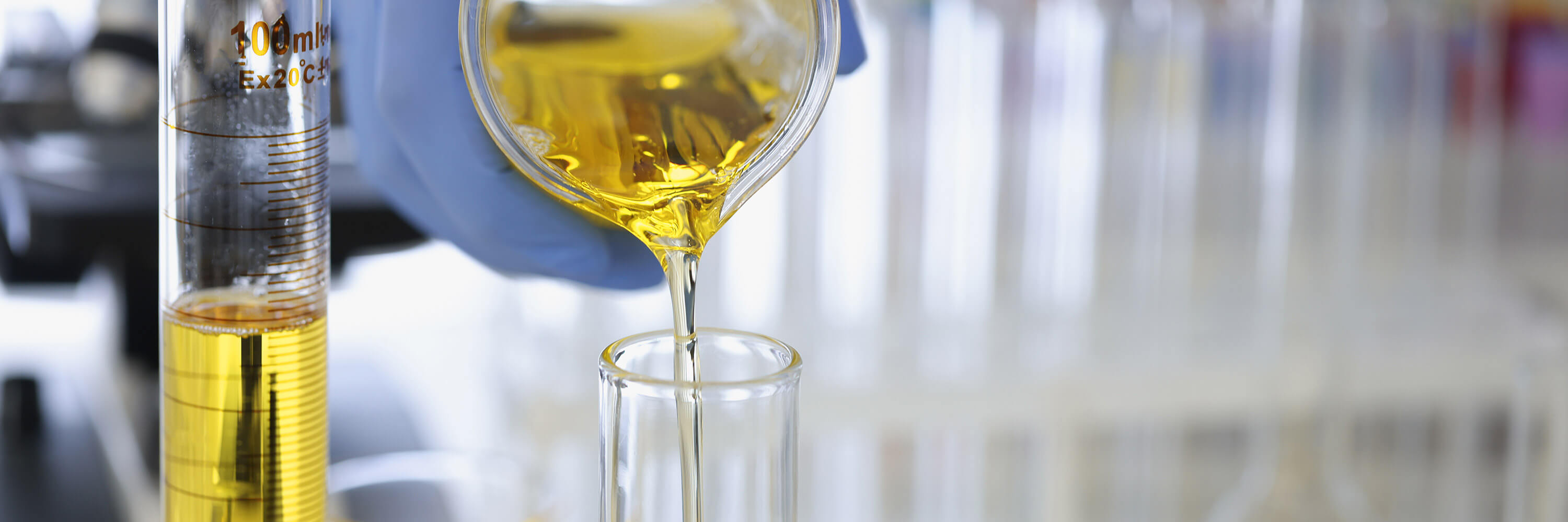 A hand in a rubber glove pours a yellow liquid into a flask in a chemical laboratory, shown in close-up as part of a pharmacological production process.