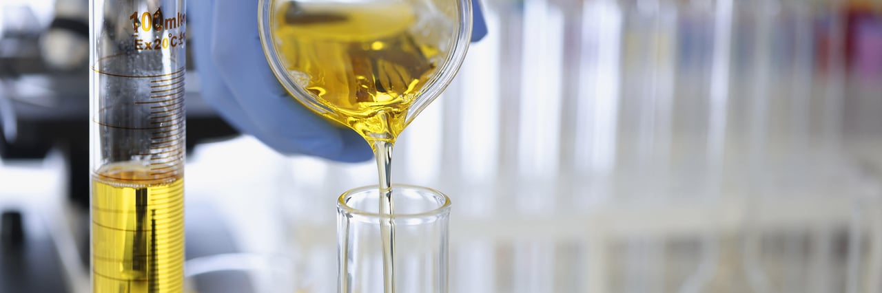 A hand in a rubber glove pours a yellow liquid into a flask in a chemical laboratory, shown in close-up as part of a pharmacological production process.