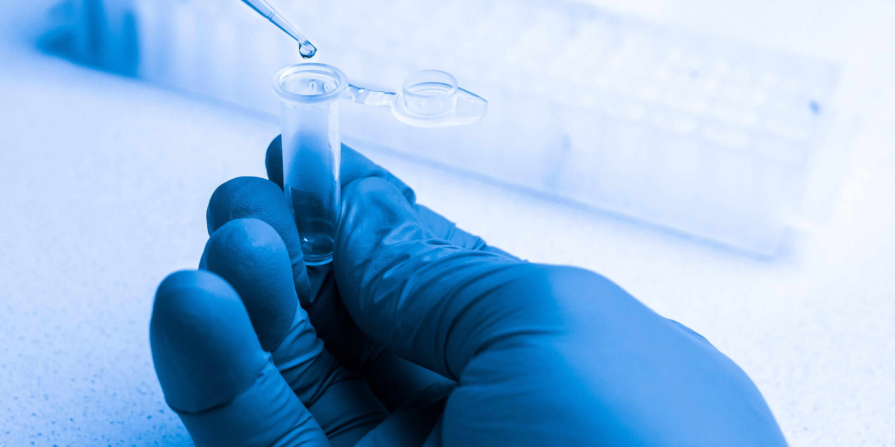 Close-up of a gloved hand holding a small sample tube while liquid is added with a pipette.