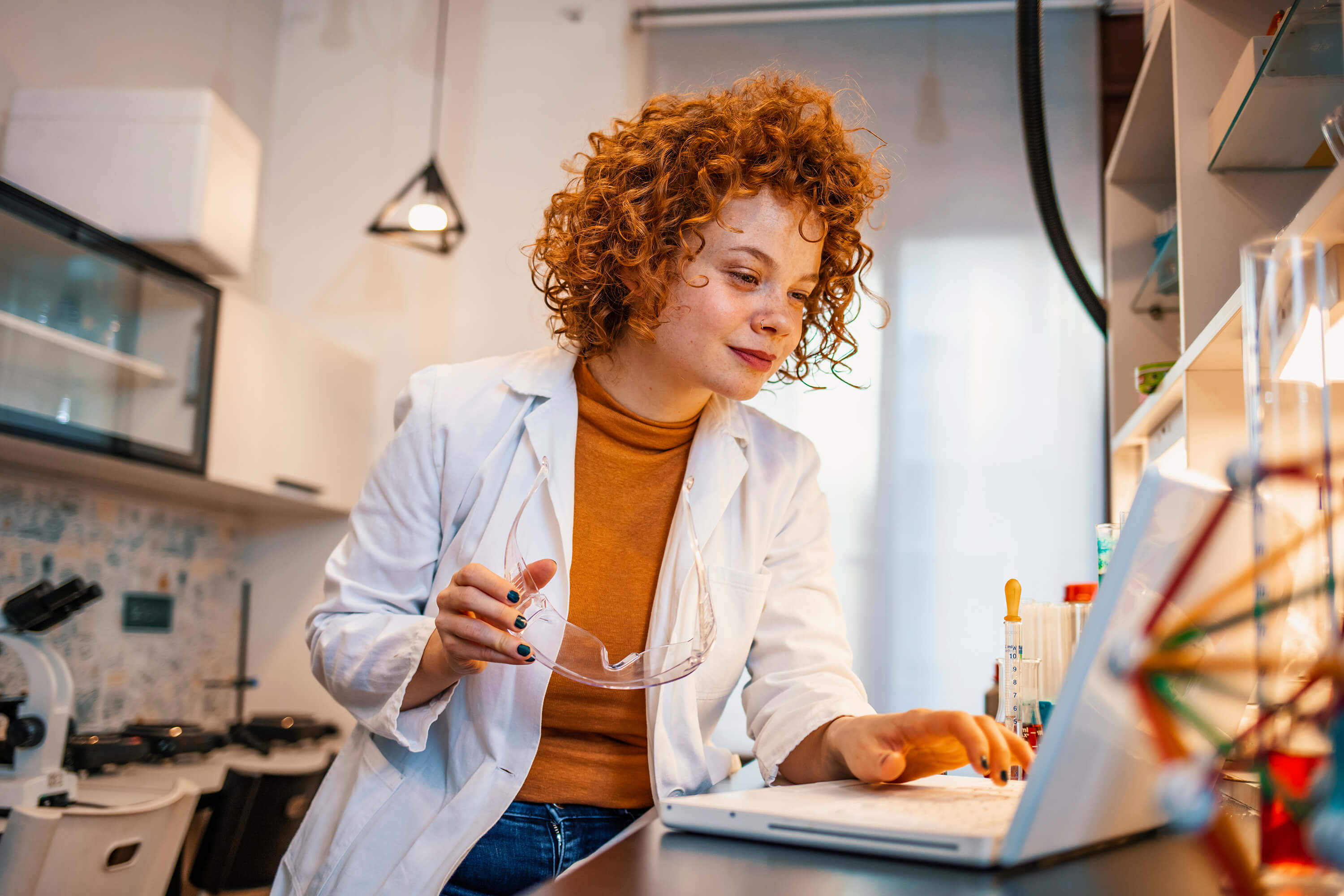 A scientist sits at a table using a laptop with a microscope beside her while reviewing test results.