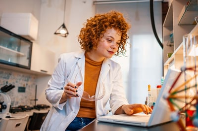 A scientist sits at a table using a laptop with a microscope beside her while reviewing test results.