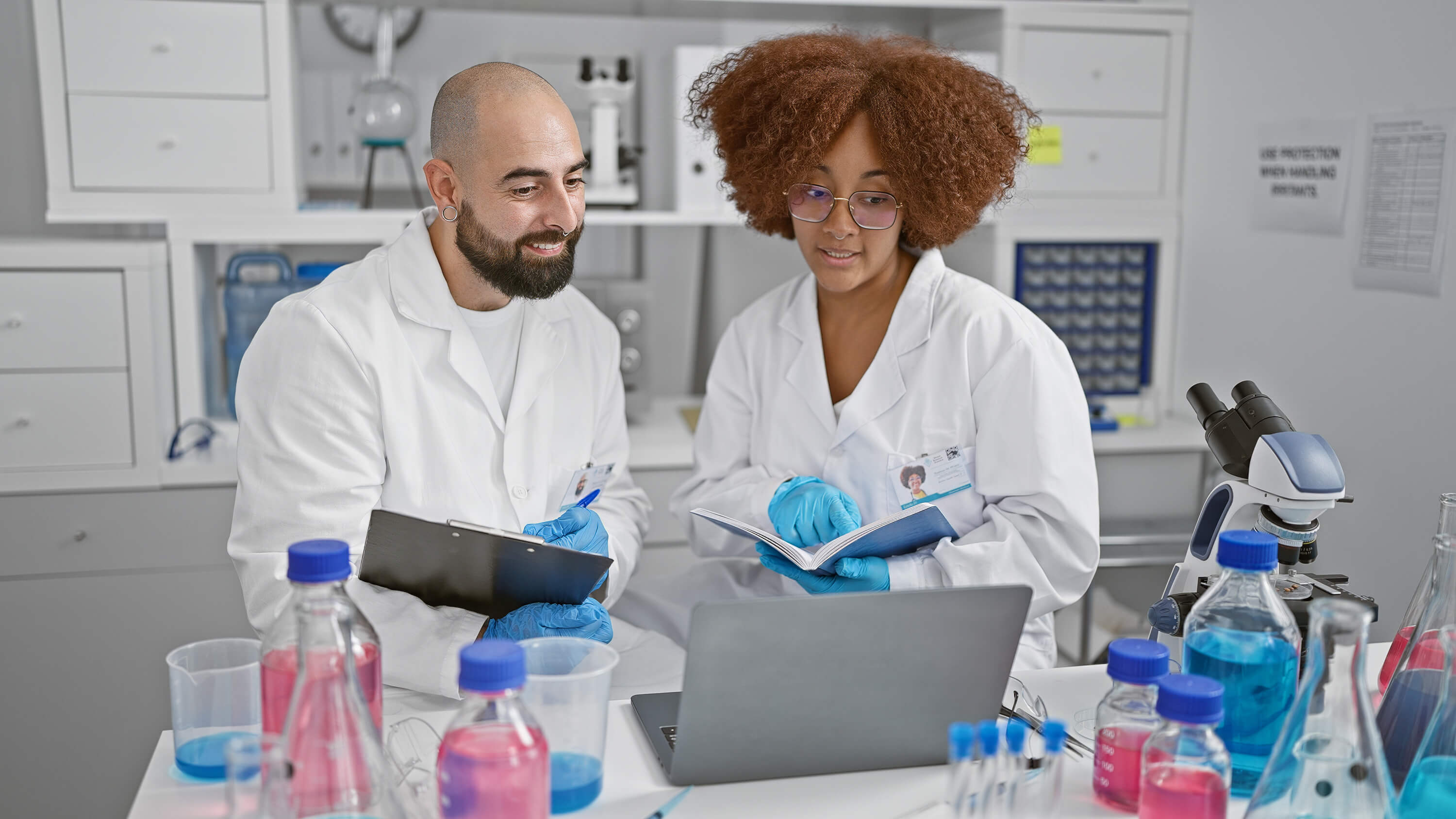 Two smiling scientists engage in online conference, reading a book and writing on clipboard in their lab while working on a medical research.