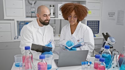 Two smiling scientists engage in online conference, reading a book and writing on clipboard in their lab while working on a medical research.