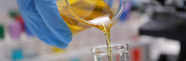 Gloved hand pouring a yellow liquid into a glass beaker in a laboratory.