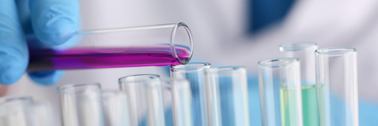 Gloved hand pouring purple liquid into test tubes in a laboratory.