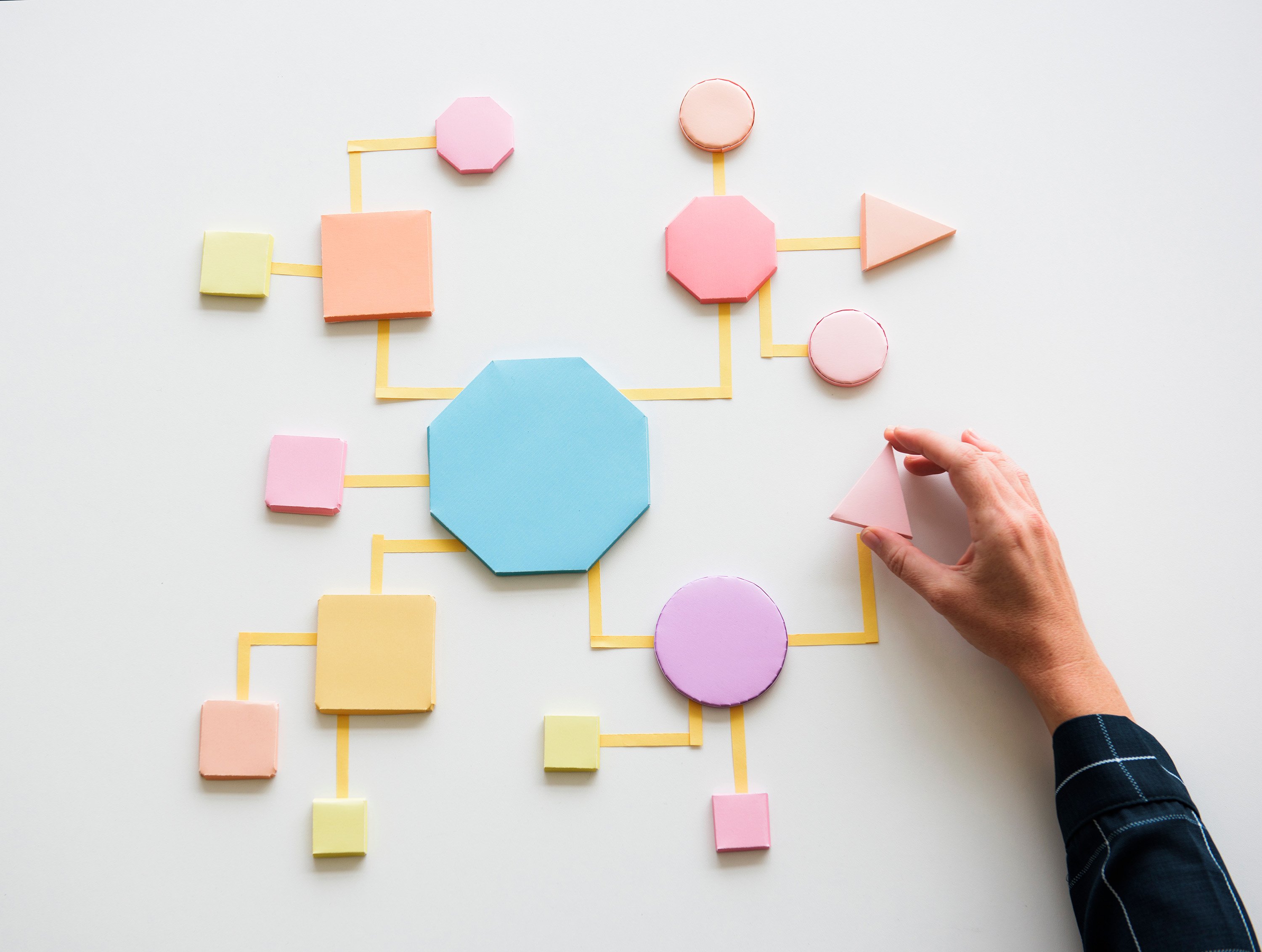 Hand placing a paper shape into a colorful business process flowchart made of connected geometric shapes on a white surface.