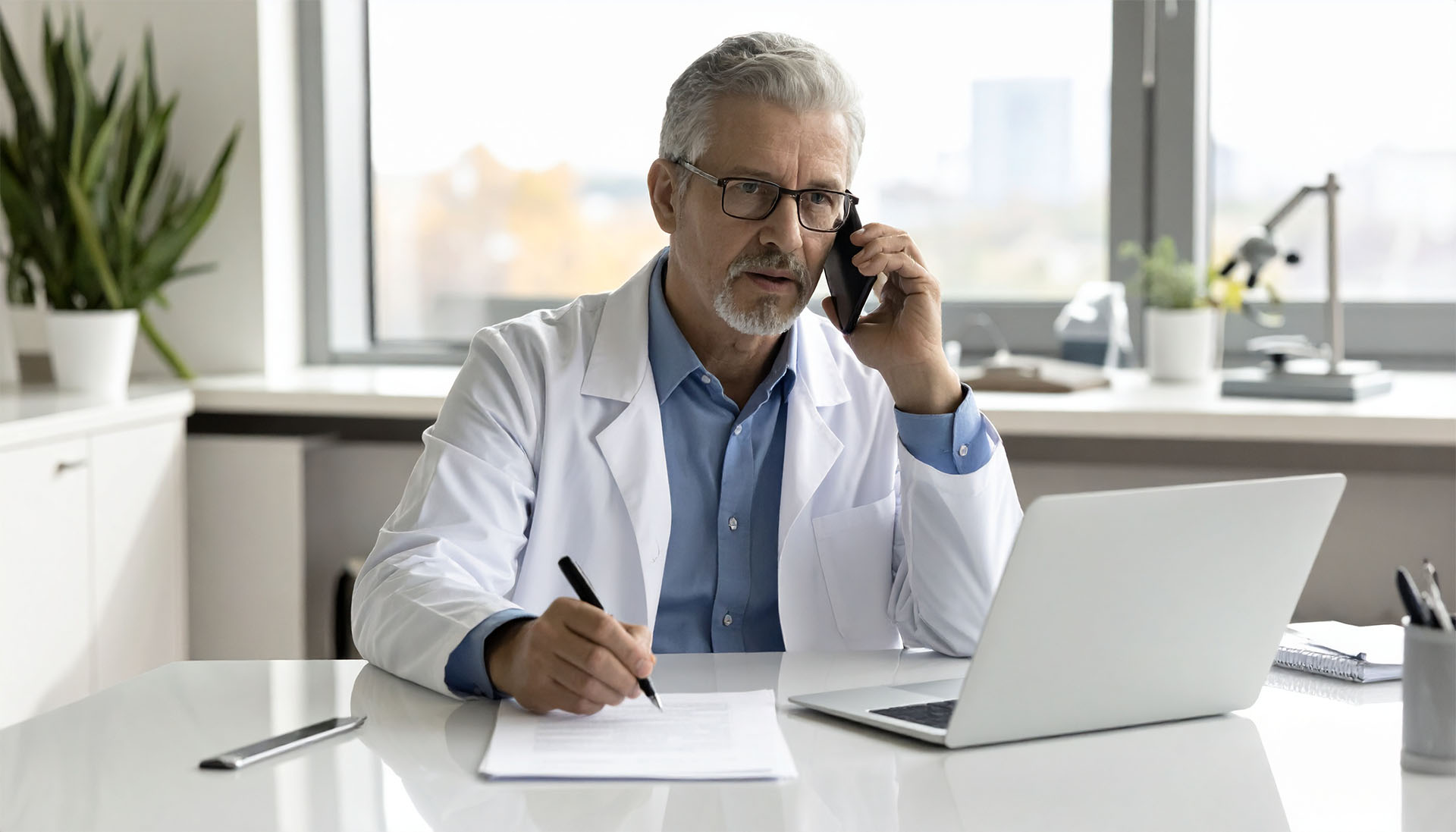 Laboratory professional using a laptop and taking notes while speaking on a phone at a desk.