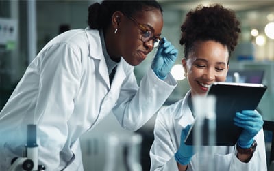 Two laboratory workers in lab coats and gloves review data on a digital tablet in a modern lab.