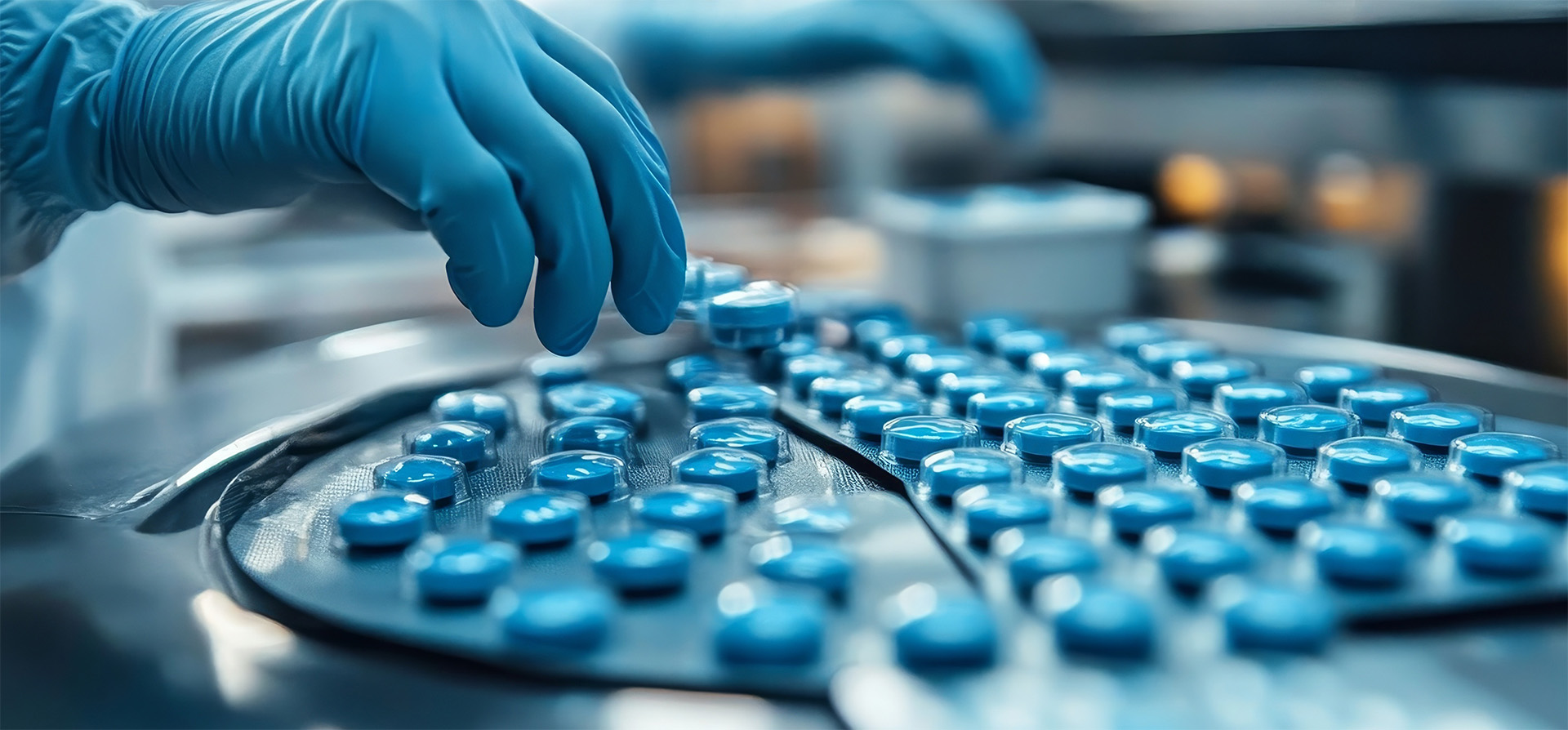 A lab worker in gloves carefully handles blue capsules on a production line, emphasizing precision and hygiene in pharmaceutical manufacturing.