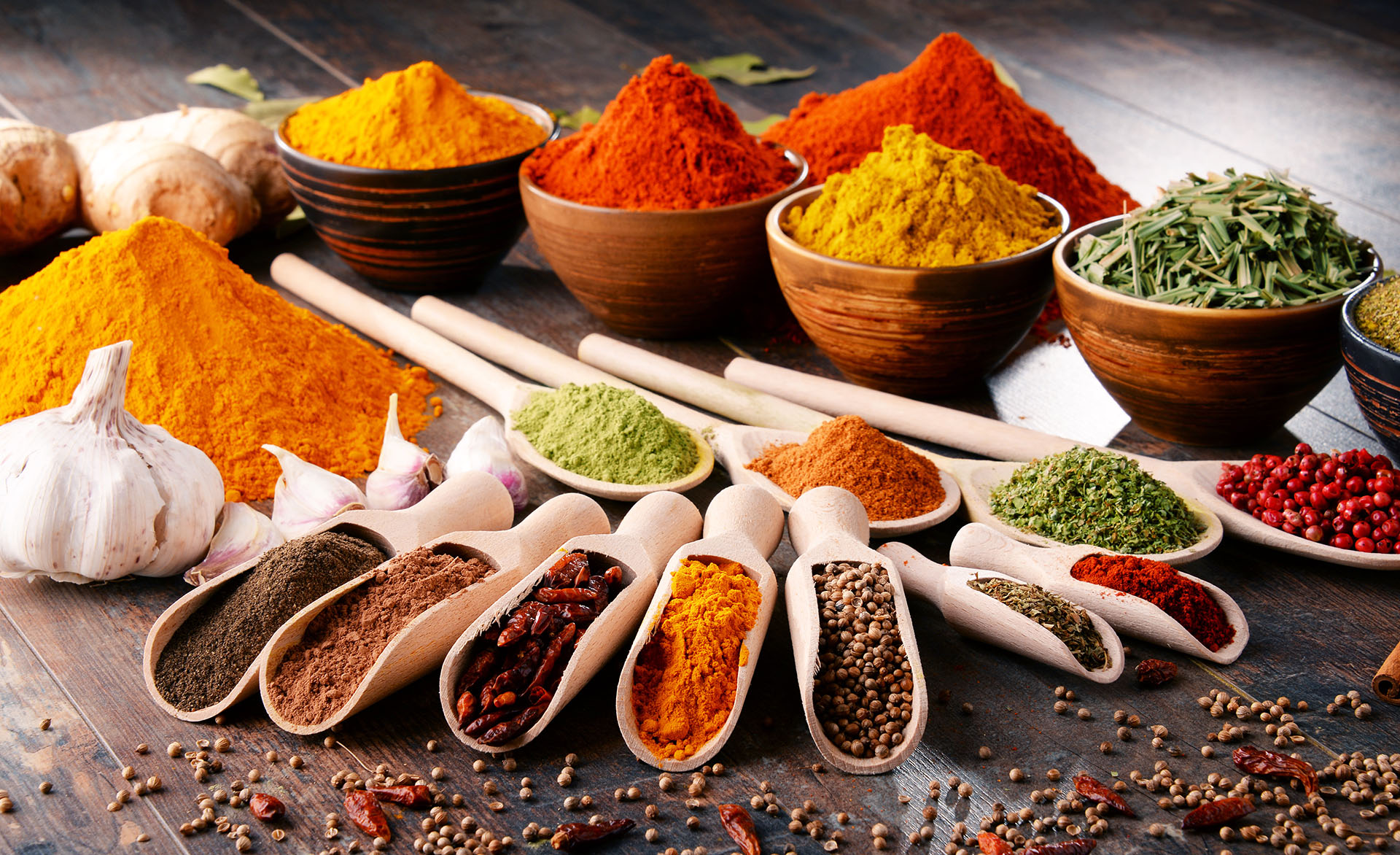 Assorted herbs & spices in bowls and wooden scoops arranged on a rustic wooden surface.