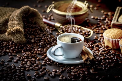 Cup of hot coffee with steam, surrounded by roasted coffee beans, cinnamon sticks, and a burlap sack.