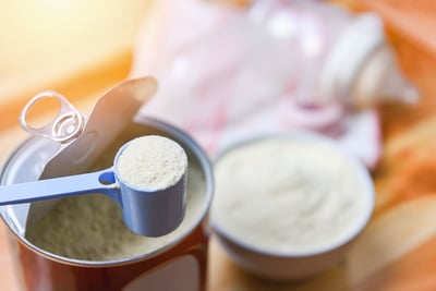 Measuring scoop with powdered milk over an open container on a wooden surface.