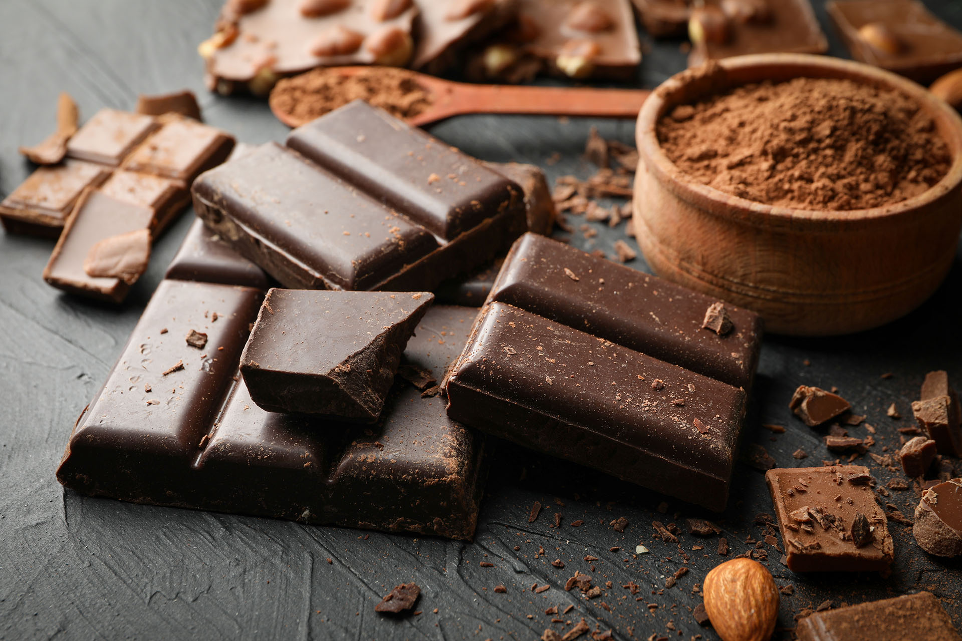 Dark chocolate pieces with cocoa powder in a wooden bowl on a dark surface.