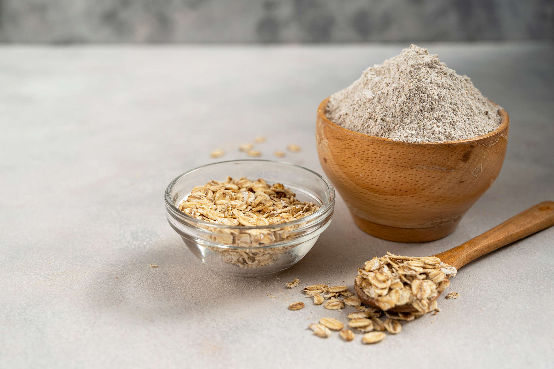 Oat flakes and oat flour in bowls with a wooden spoon on a neutral surface.
