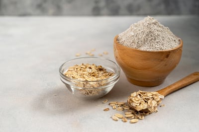 Oat flakes and oat flour in bowls with a wooden spoon on a neutral surface.