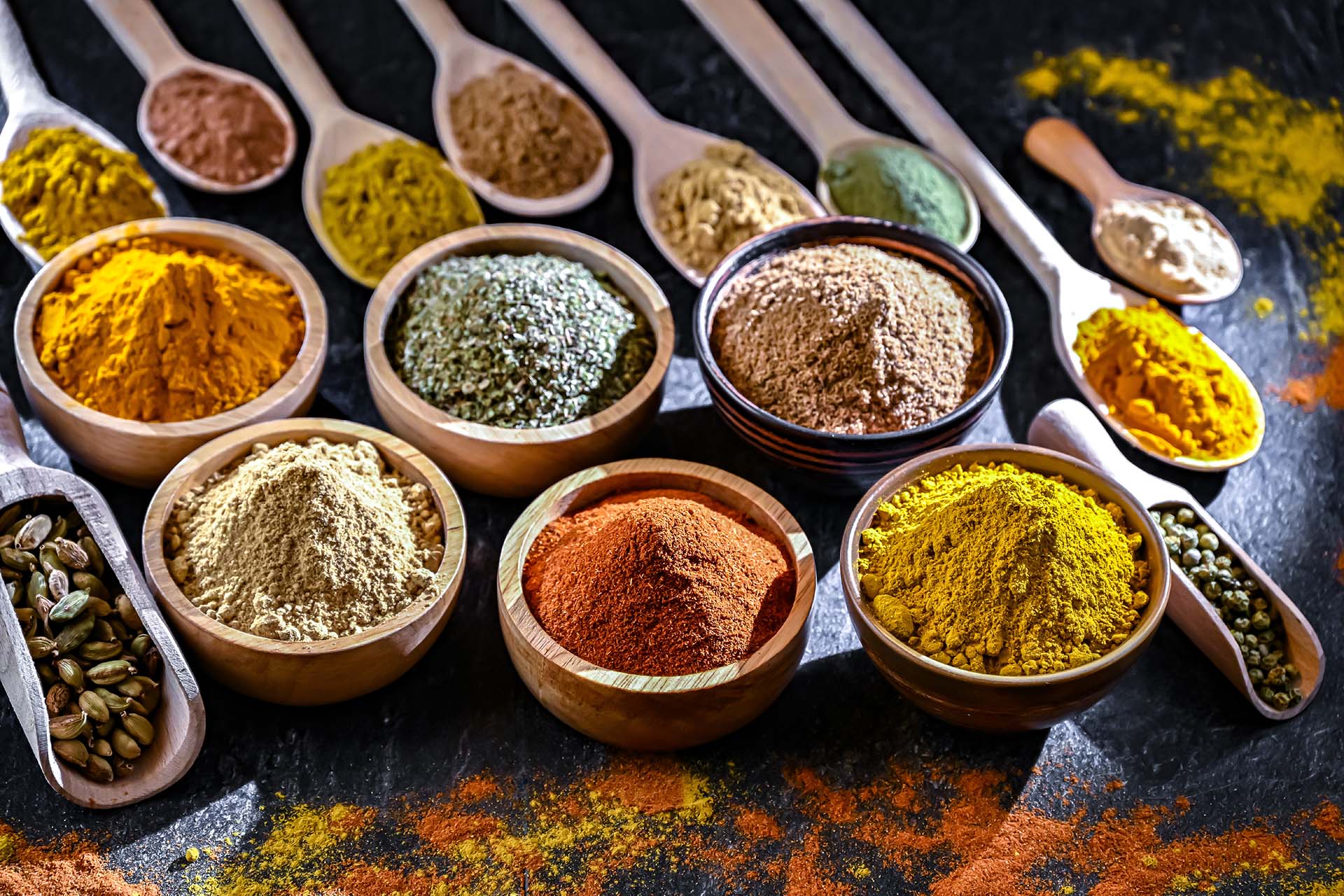 Assorted ground spices in wooden bowls and spoons arranged on a dark surface.